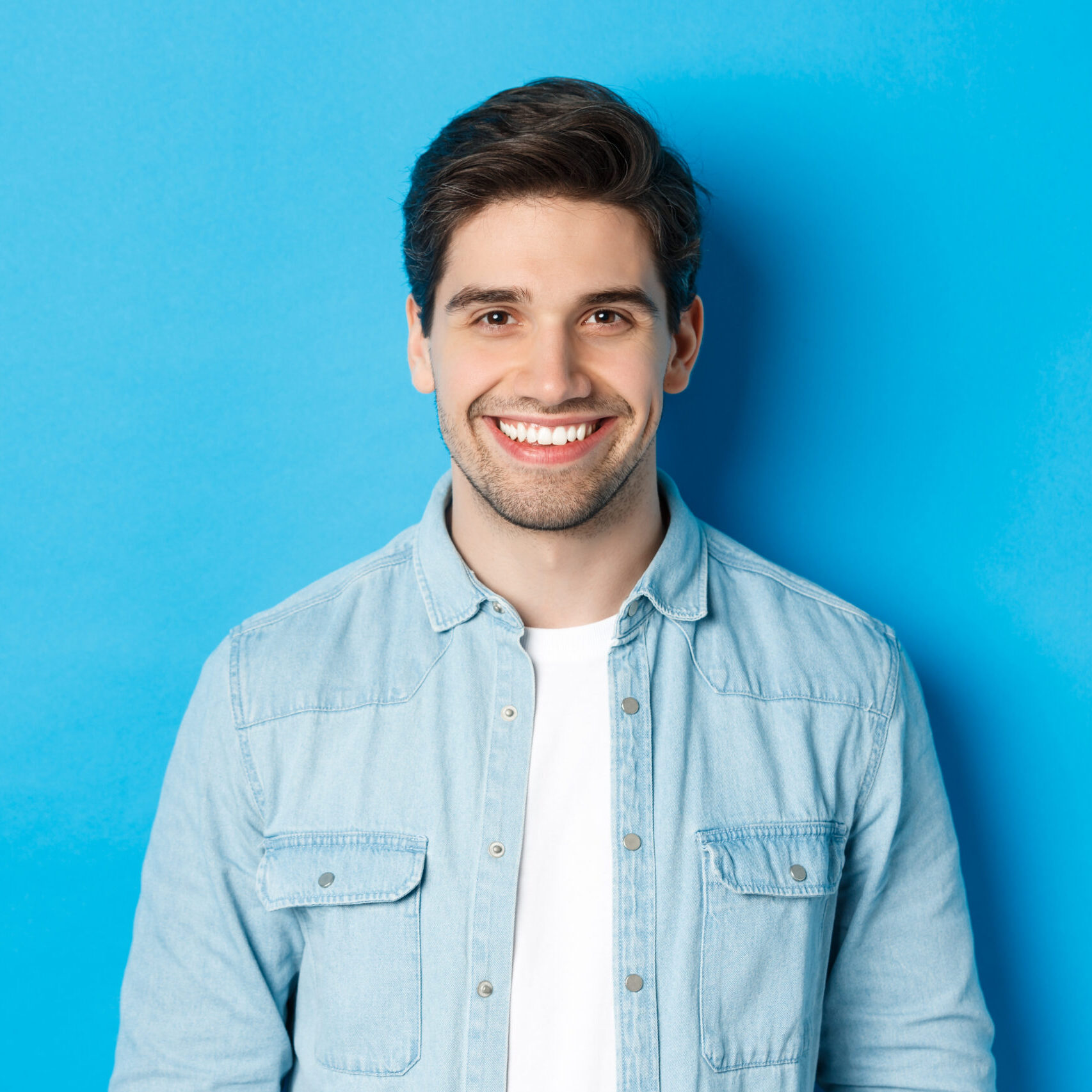 Close-up of young successful man smiling at camera, standing in casual outfit against blue background.